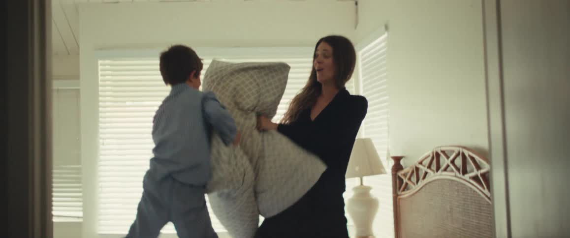 A happy mother and her son enjoy a playful pillow fight in slow motion on a bed in a sunlit bedroom