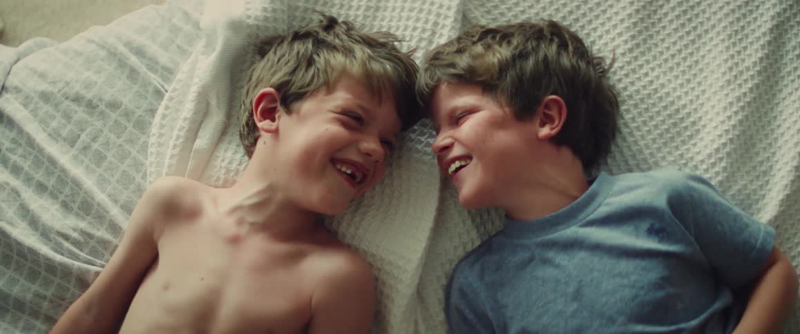 Two smiling boys lie face to face on a bed sharing a playful, intimate moment indoors