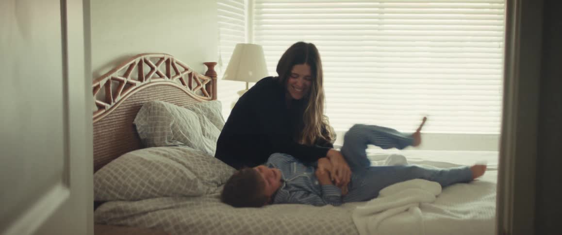 Mother playfully tickles laughing child in striped pajamas on a sunlit bedroom bed