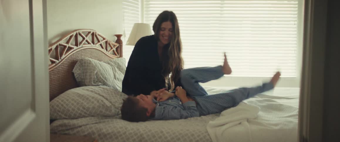 Mother playfully tickles laughing child in striped pajamas on a sunlit bedroom bed