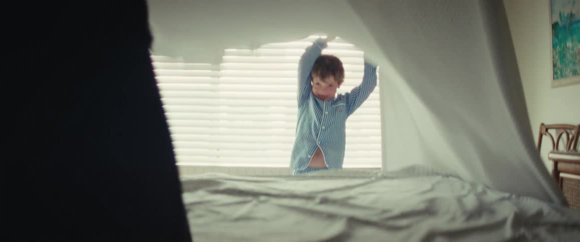 A happy little boy in pajamas plays peek-a-boo with a white blanket as his mom makes the bed