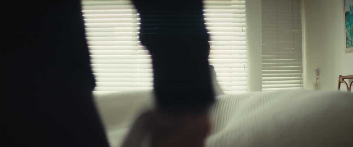 A happy little boy in pajamas plays peek-a-boo with a white blanket as his mom makes the bed