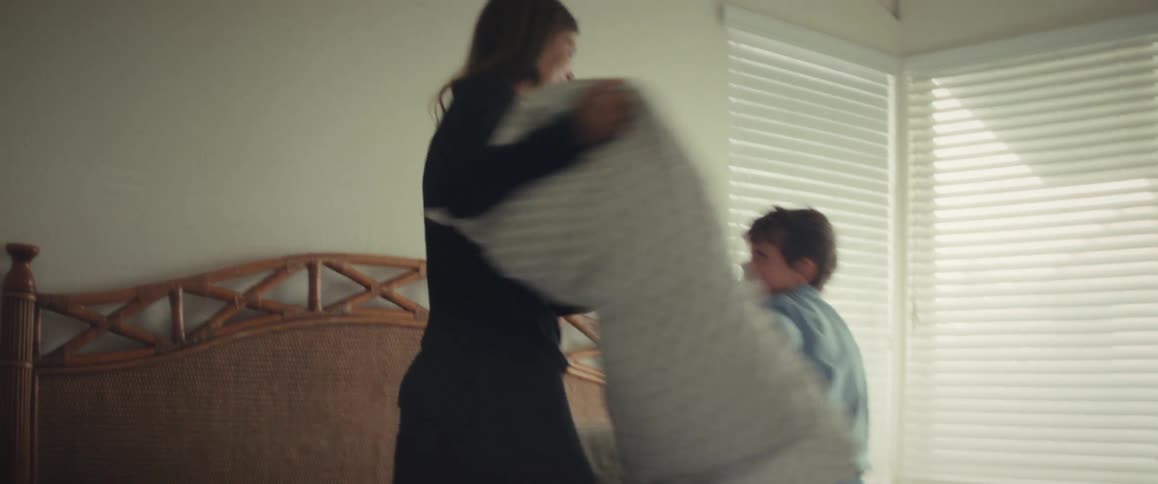 A joyful mother and her young son laugh while having a pillow fight on the bed in slow motion