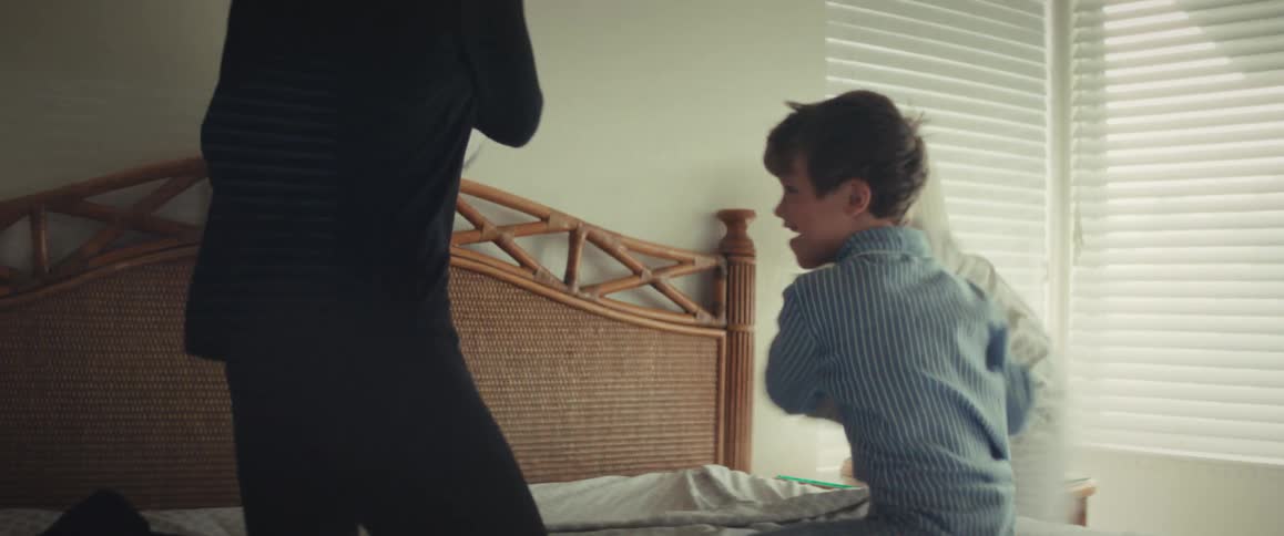 A joyful mother and her young son laugh while having a pillow fight on the bed in slow motion