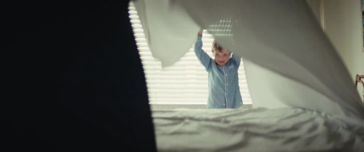 A happy boy in pajamas plays peek-a-boo with a parent on a sunlit bed