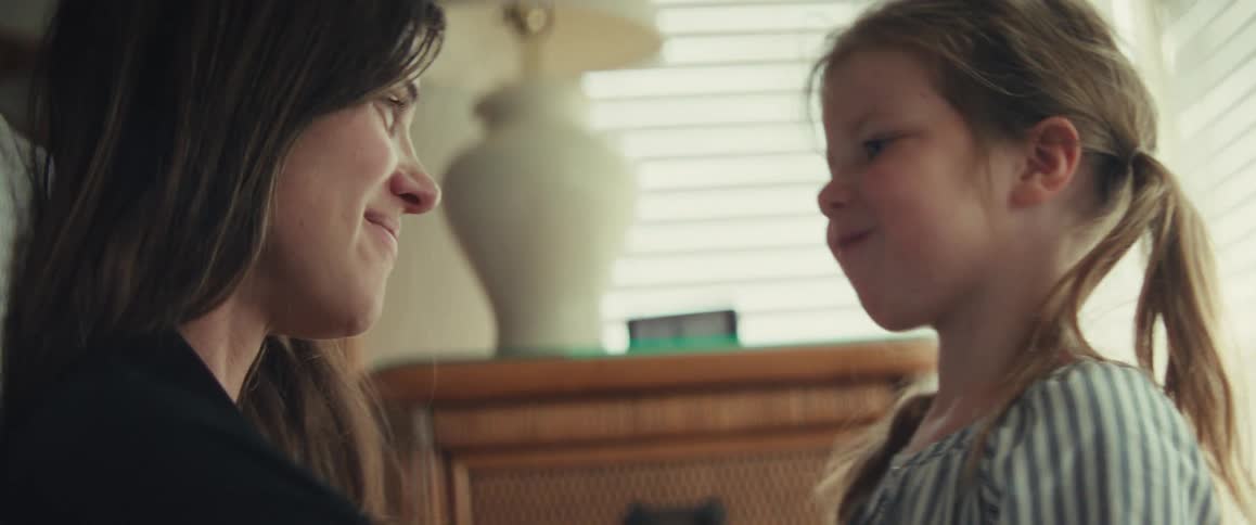 A mother and her young daughter share a playful and affectionate kiss in a softly lit room