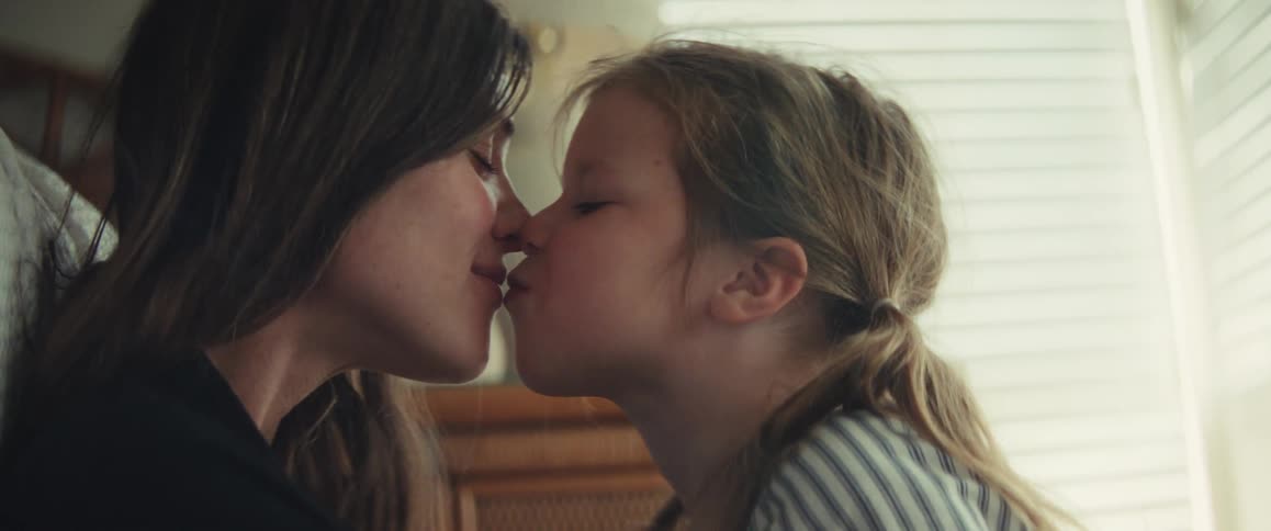 A mother and her young daughter share a playful and affectionate kiss in a softly lit room