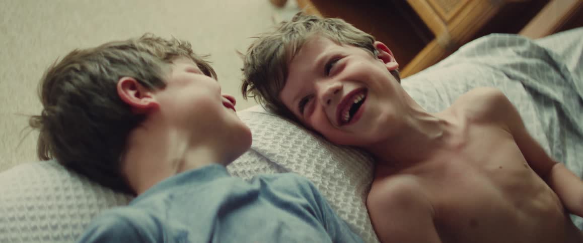 Two young brothers lie on pillows indoors smiling and laughing together in a cozy bedroom