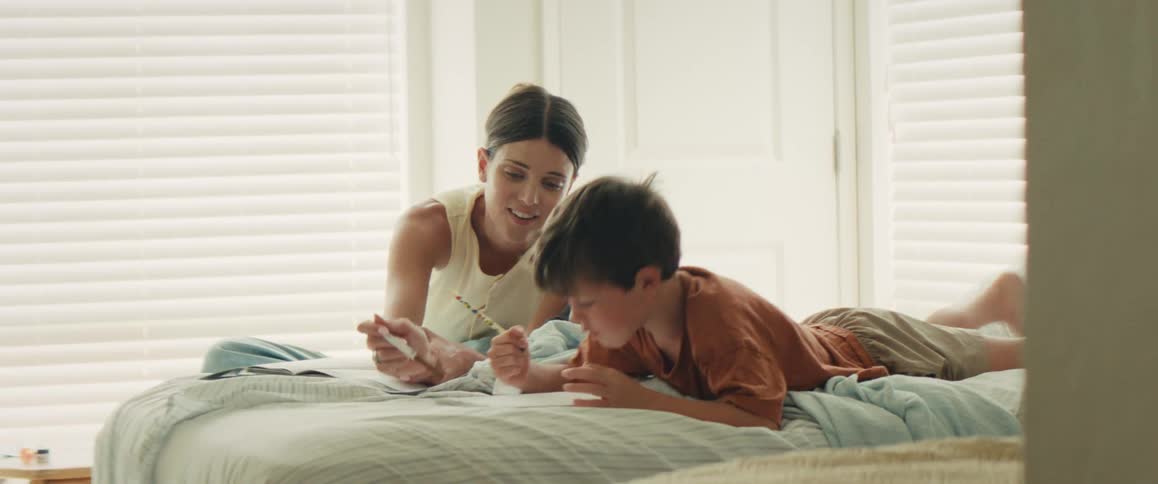 A smiling mother helps her young son with his homework on the bed, and they celebrate with a high-five