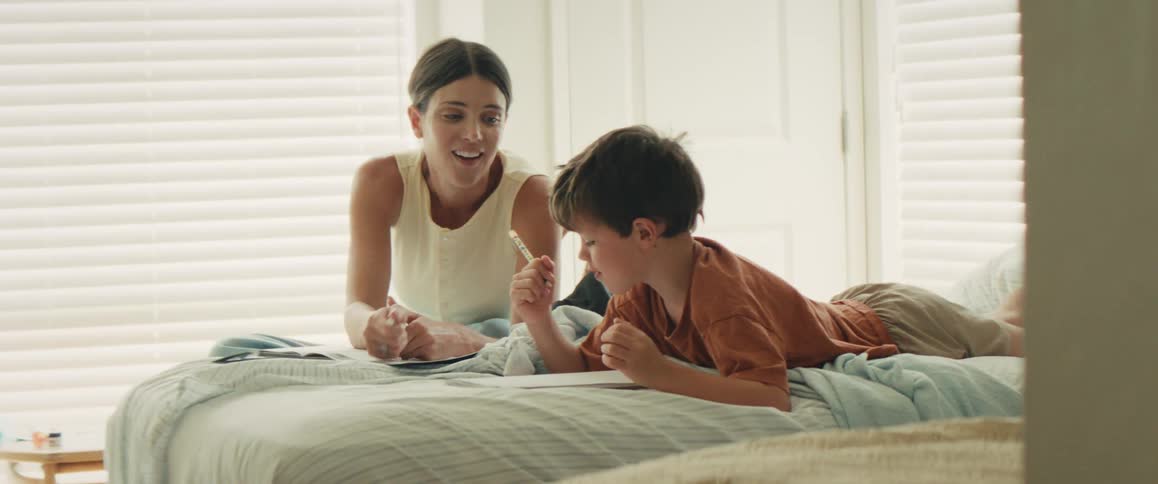 A smiling mother helps her young son with his homework on the bed, and they celebrate with a high-five
