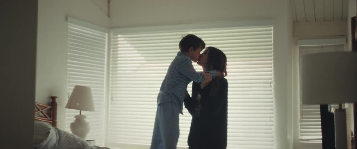 A young boy in pajamas stands on the bed to give his mother a sweet kiss and a long, loving hug in a brightly lit bedroom