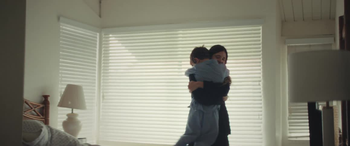 A young boy in pajamas stands on the bed to give his mother a sweet kiss and a long, loving hug in a brightly lit bedroom