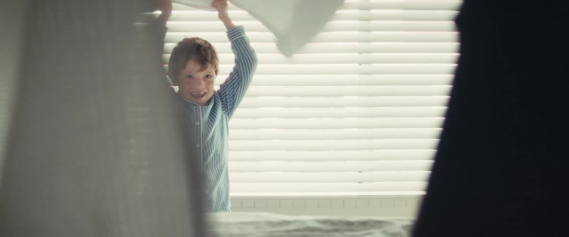A playful young boy in pajamas laughs while playing peek-a-boo with a white blanket in a sunlit bedroom