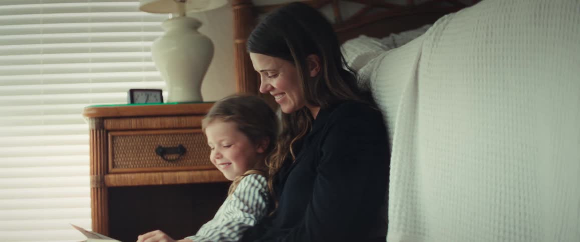 A mother and daughter smile and laugh while reading a storybook together on the floor of a sunlit bedroom