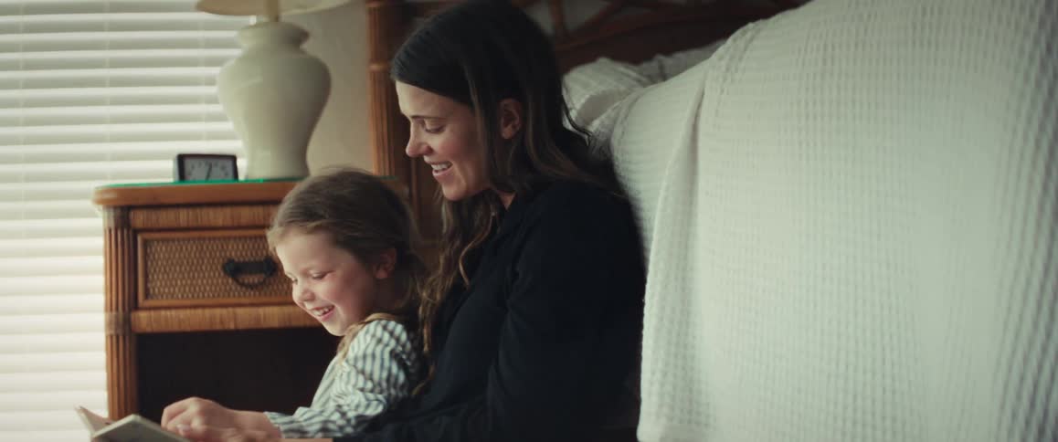 A mother and daughter smile and laugh while reading a storybook together on the floor of a sunlit bedroom