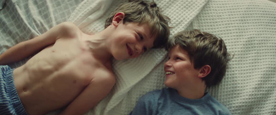 Two smiling boys lie on a white bed sharing a playful affectionate moment at home