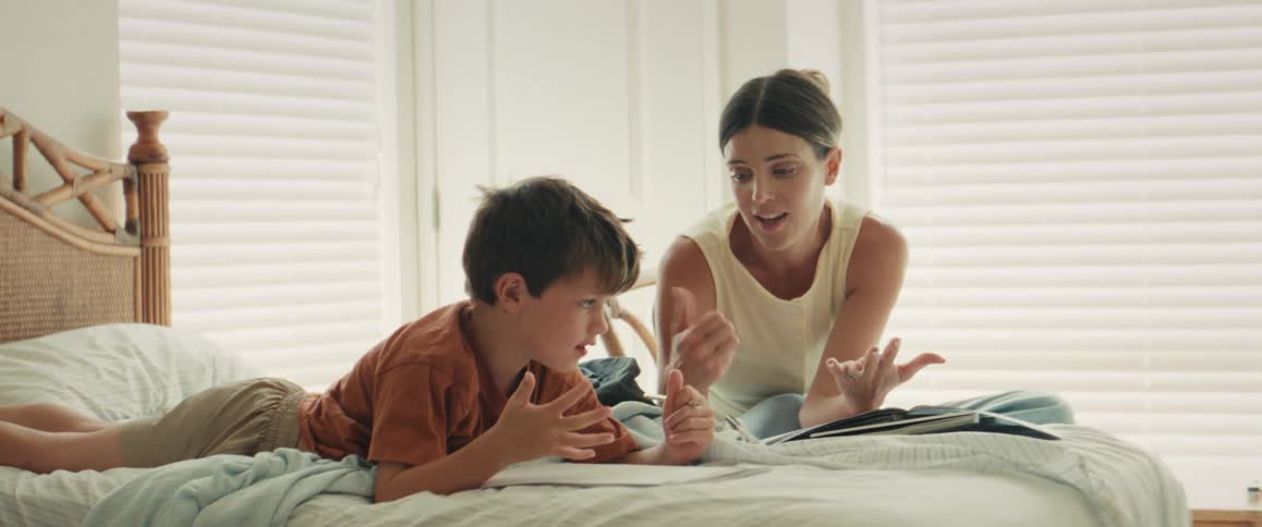 A caring mother helps her young son with his homework, patiently teaching him to count as they sit together on a sunlit bed