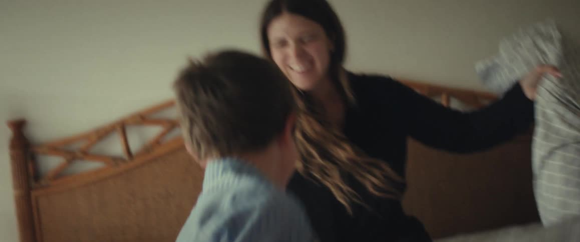 A young boy in pajamas has a fun pillow fight with his smiling mother on a bed in slow motion