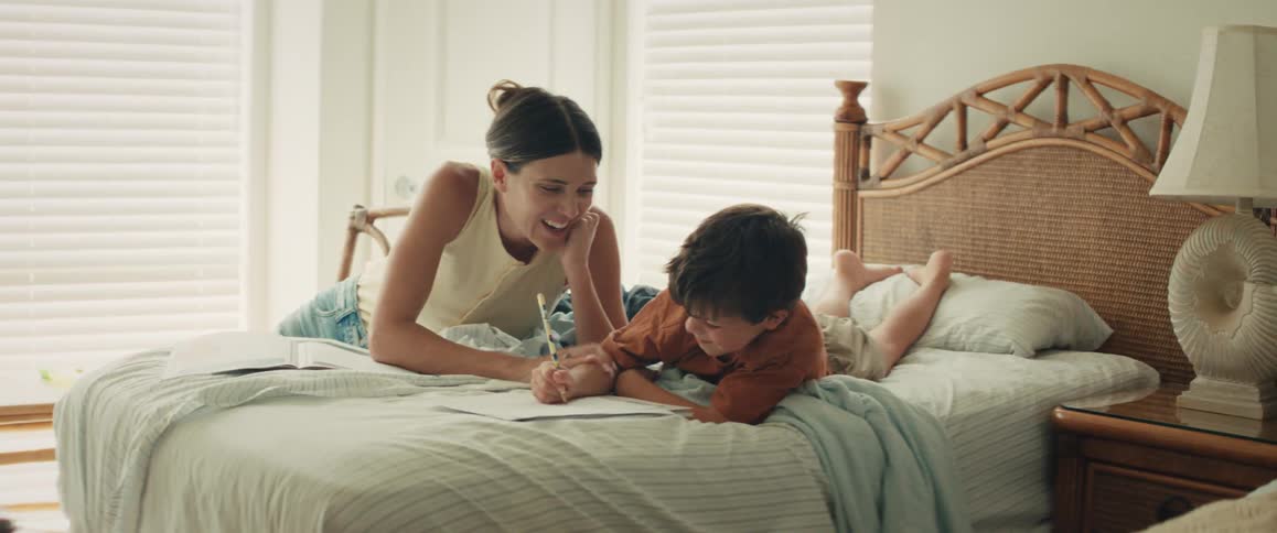 A loving mother smiles as she helps her young son draw while they relax on a bed together in a sunlit bedroom