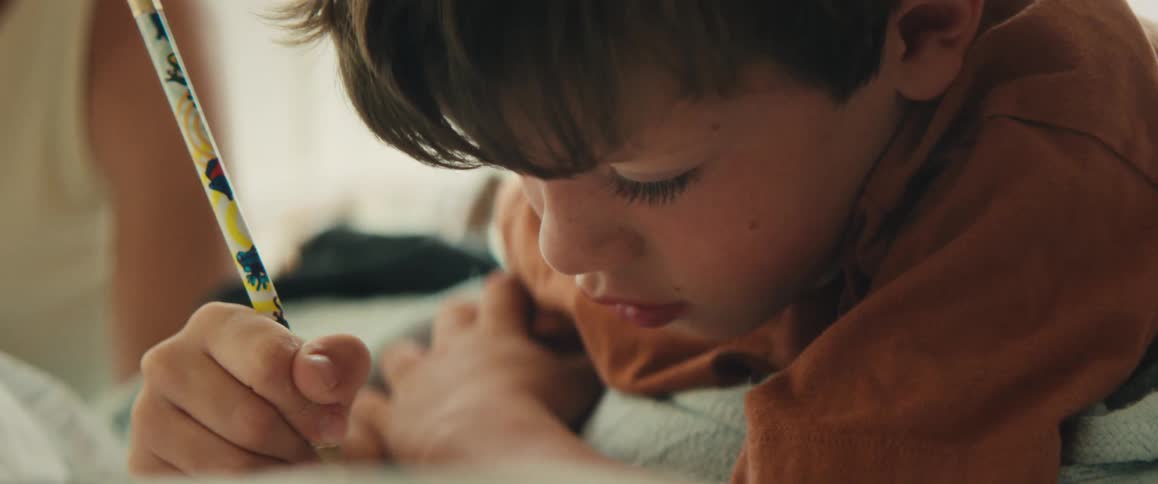 A young boy with brown hair intensely concentrates while drawing with a pencil in a close-up shot