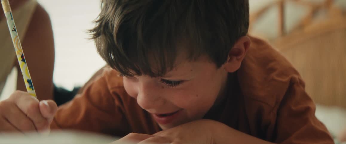 Smiling young boy lies on the floor drawing with a pencil at home in warm natural light