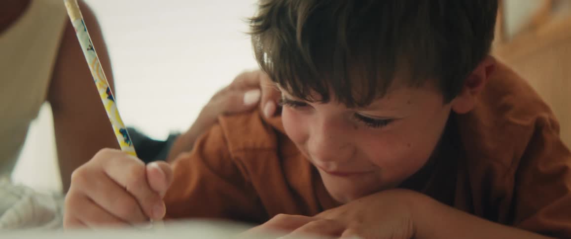 Smiling young boy lies on the floor drawing with a pencil at home in warm natural light