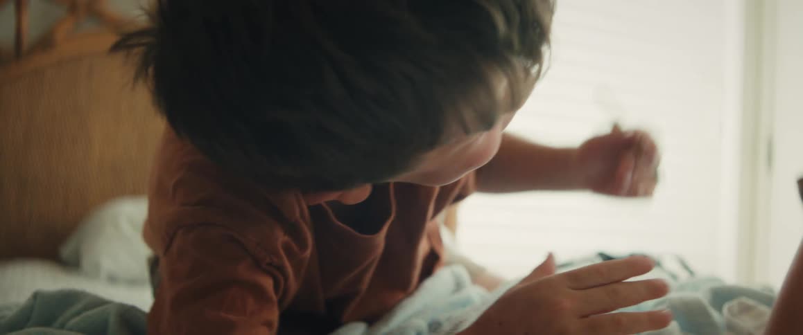 A focused young boy lies on his bed in a sunlit room, intently drawing with a colored pencil