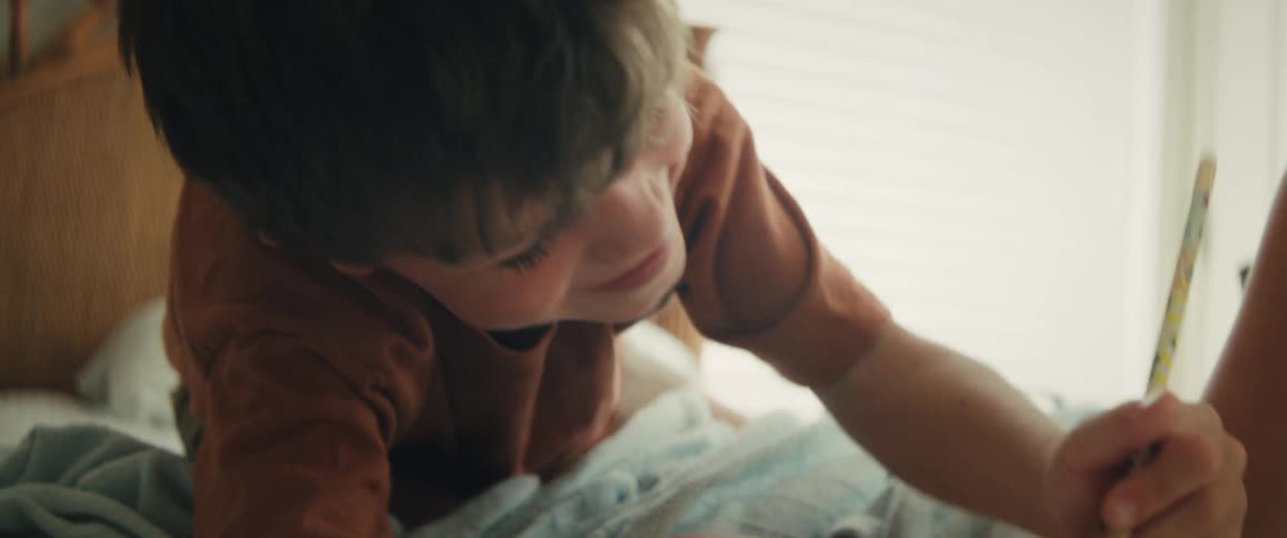 A focused young boy lies on his bed in a sunlit room, intently drawing with a colored pencil