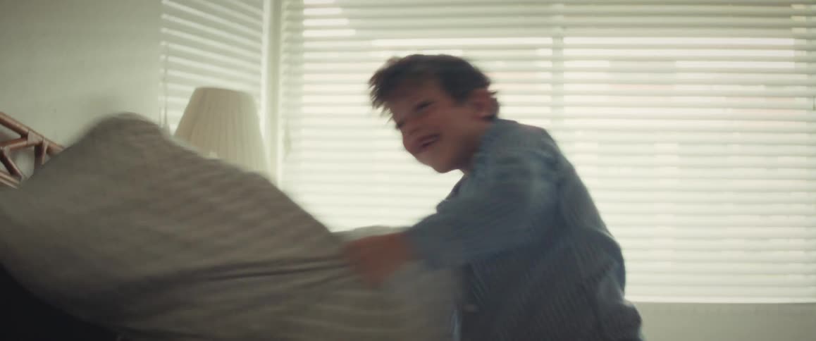 A joyful mother and her young son laugh and have a playful pillow fight in their pajamas on a bed in slow motion