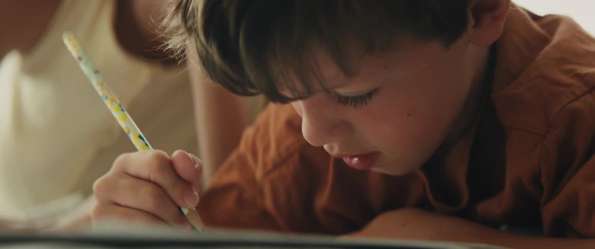 A close-up of a young boy concentrating on drawing with a pencil while his mother helps him