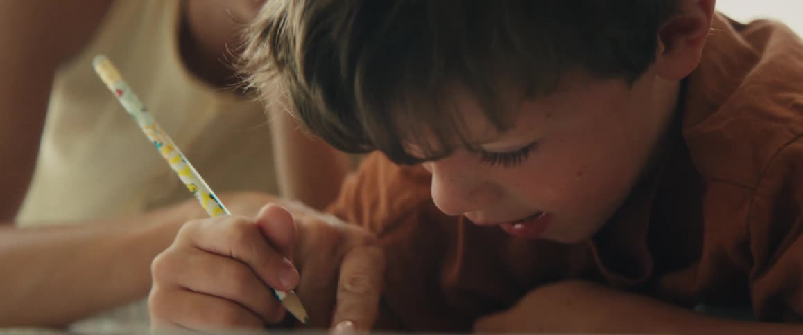 A close-up of a young boy concentrating on drawing with a pencil while his mother helps him