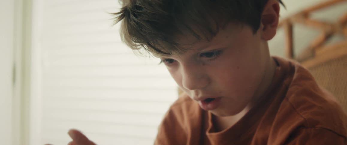 A focused young boy is seen in an intimate close-up as he concentrates on something off-screen, possibly learning or playing a game