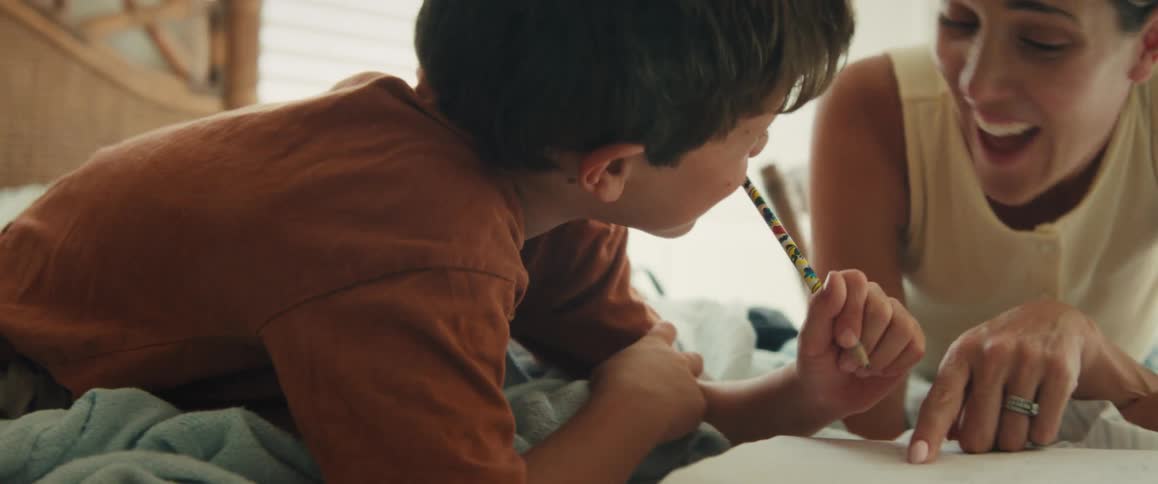 A smiling mother patiently helps her young son with his homework as they lie on a bed together in a cozy bedroom
