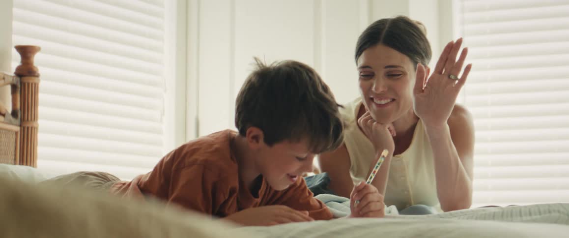 A loving mother and her young son share a happy moment drawing and laughing together while lying on a bed in a bright bedroom