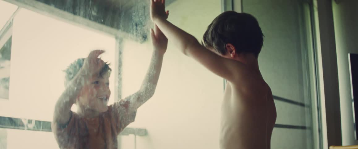 Young boy playfully presses hands against a foggy glass door indoors with warm natural light