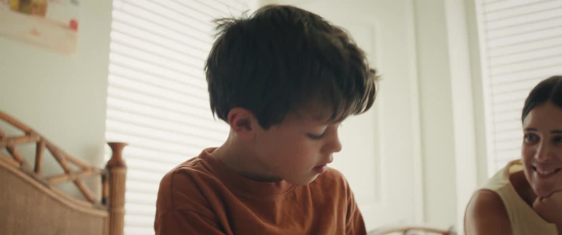 A young boy intently draws at a table while his proud mother lovingly watches over him in a brightly lit room