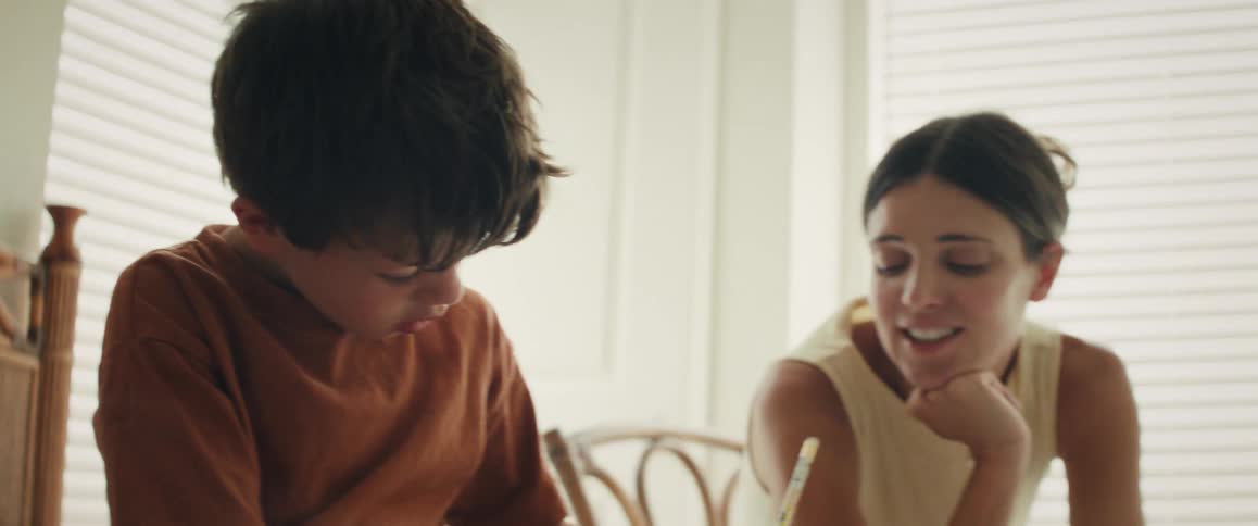 A young boy intently draws at a table while his proud mother lovingly watches over him in a brightly lit room