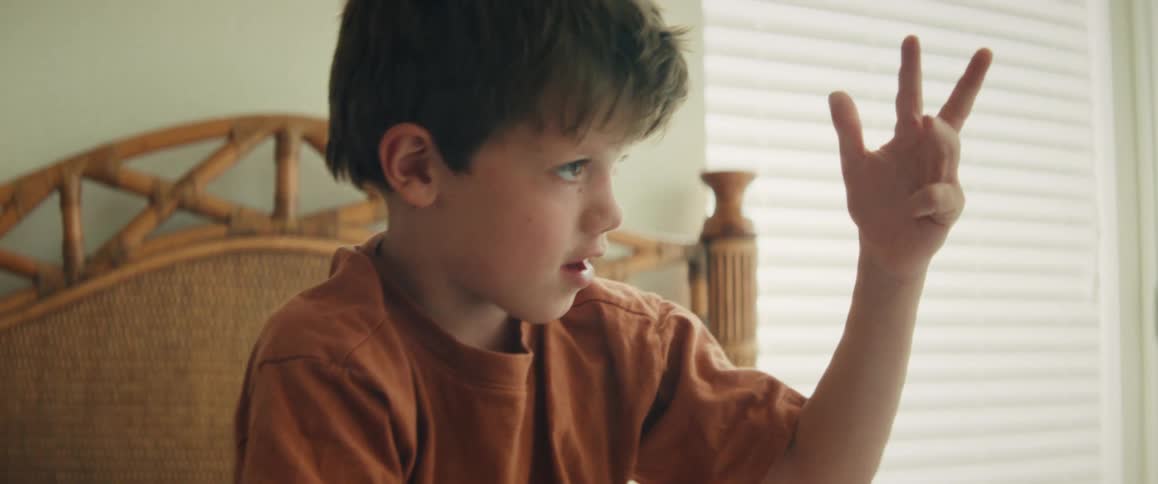 A young boy in a sunlit bedroom sits on his bed and carefully counts on his fingers