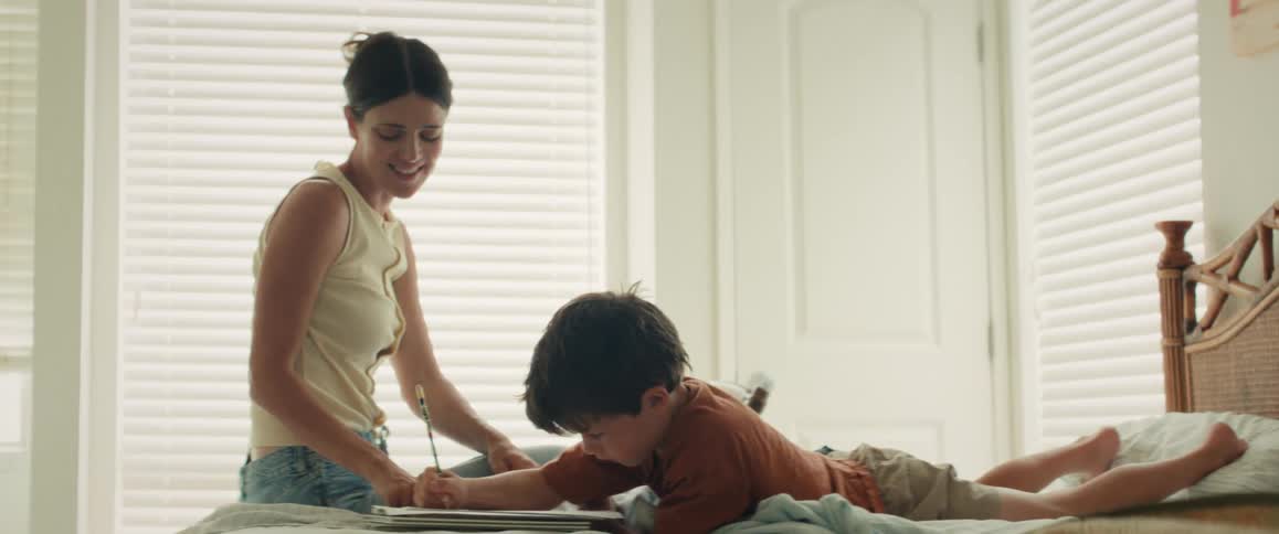 A smiling mother helps her young son draw in a notebook as they relax together on a bed in a sunlit room
