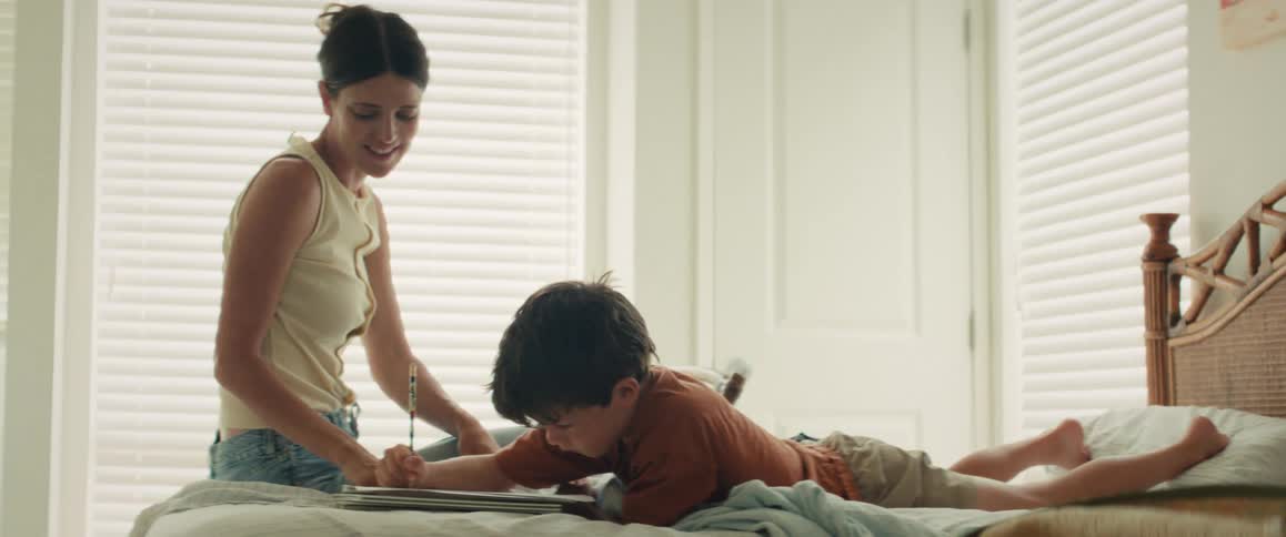 A smiling mother helps her young son draw in a notebook as they relax together on a bed in a sunlit room