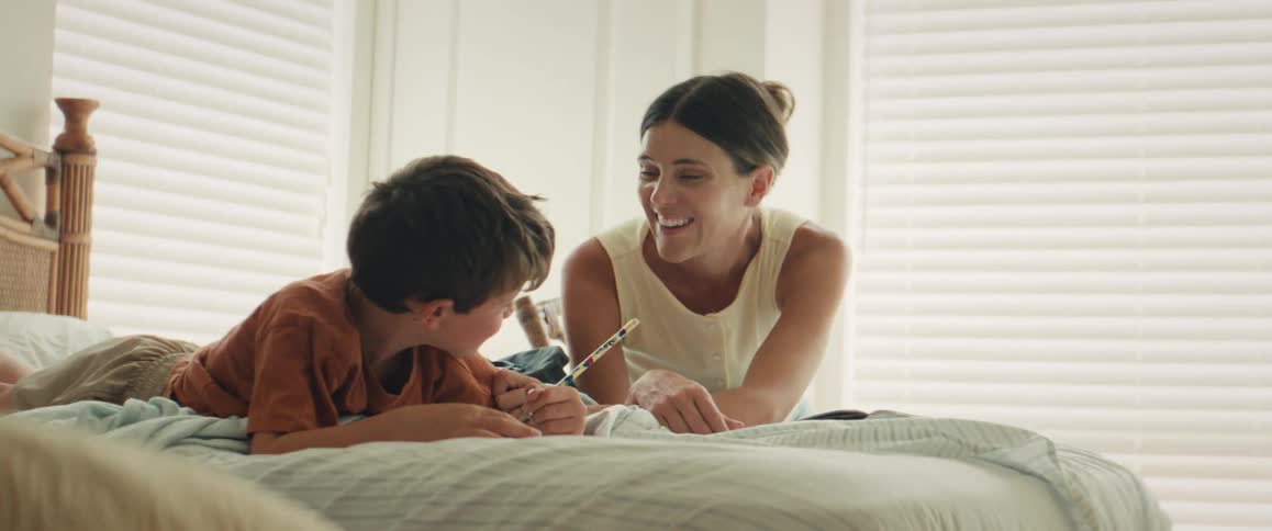 A smiling mother lovingly watches as her young son draws in a notebook while they relax on a bed in a sunlit bedroom