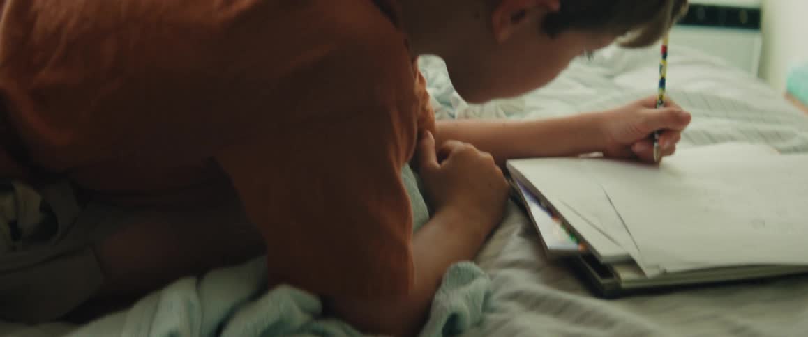 A young boy intently draws in his sketchbook while lying on his bed as his mother watches with a smile