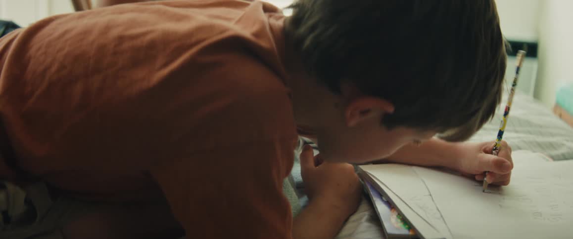 A young boy intently draws in his sketchbook while lying on his bed as his mother watches with a smile