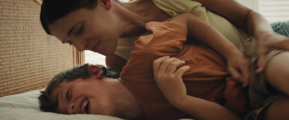 A close-up slow-motion shot shows a young boy laughing uncontrollably while his mother playfully tickles and kisses him on the bed