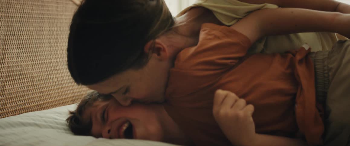 A close-up slow-motion shot shows a young boy laughing uncontrollably while his mother playfully tickles and kisses him on the bed