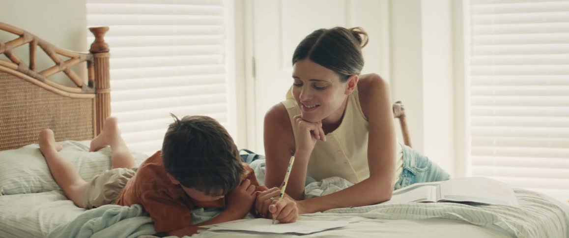 A caring mother helps her young son with his drawing as they spend quality time together on a bed in a bright, sunlit bedroom
