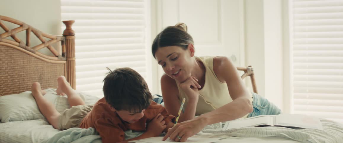 A caring mother helps her young son with his drawing as they spend quality time together on a bed in a bright, sunlit bedroom