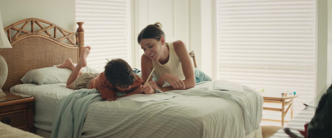 A smiling mother helps her young son draw while they relax on a bed, sharing a high-five and a loving kiss in a bright bedroom