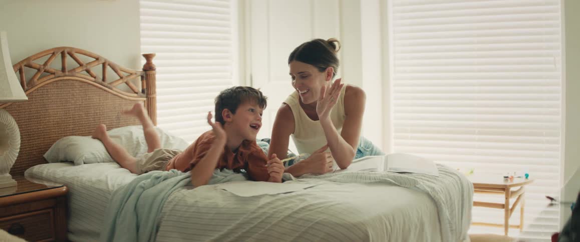 A smiling mother helps her young son draw while they relax on a bed, sharing a high-five and a loving kiss in a bright bedroom
