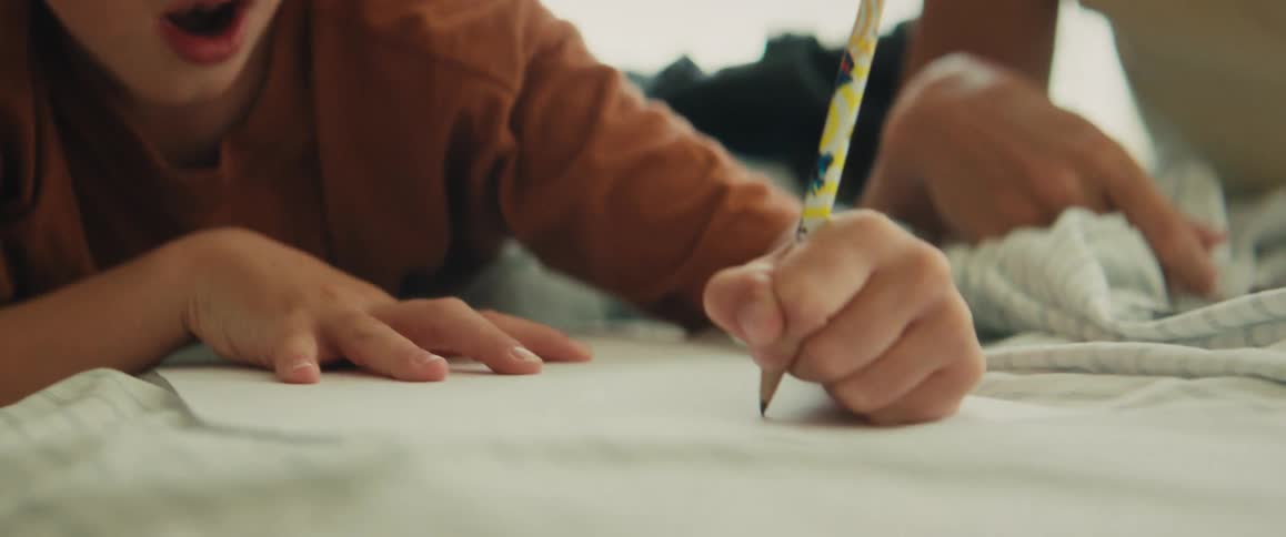 A focused young boy lies on his stomach, concentrating intently as he draws with a pencil on a piece of paper